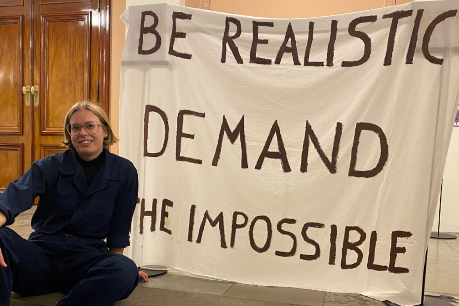 Pinja Vuorinen, iklädd en blå halare, sitter bredvid en banderoll med texten "Be Realistic, demand the impossible".