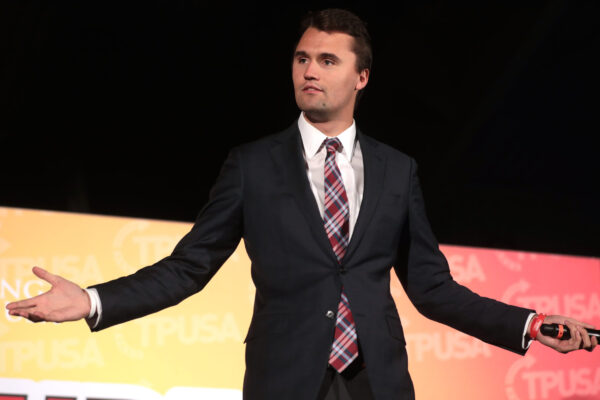 Charlie Kirk iklädd svart kostym. Han står på en sen med händerna vid sidan på ett frågande sätt.