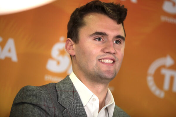 Charlie Kirk framför orange bakgrund. Han har på sig en grå kostym och ler.
