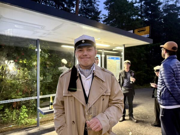 En person står framför en busshållplats med ett glas skumvin i handen. Hen är iklädd en studentmössa med lång tofs. Bakgrunden lyses upp av hållplatsens belysning, men bakom hållplatsen är det mörkt.