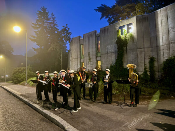 En grupp personer spelar olika instrument. De är iklädda vita studentmössor med långa svarta tofsar och fracker. De står framför ett grått hus som har en upplyst text: "TF". Det är mörkt, bara gatubelysningen lyser upp musikanterna.