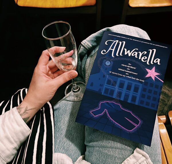 En hand som håller i ett skumvinsglas. Bredvid glaset ligger ett programblad i personens knä. Programbladet är mörkblått med "Allwarella skrivet i vitt.