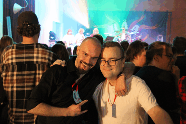 Ville Anttinen och Dennis Granith tittar leende in i kameran. Ville Anttinen håller ut tungan. I bakgrunden syns en publik som står vända mot en scen med grönt ljus och strålkastare.