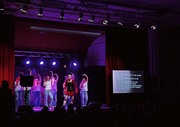 En person i rödglittrig klänning står på scen. Bakom hen dansar bakgrundsdansare. Scenen är upplyst med lila lampor. bredvid scenen finns en skärm där låttexten projiceras.