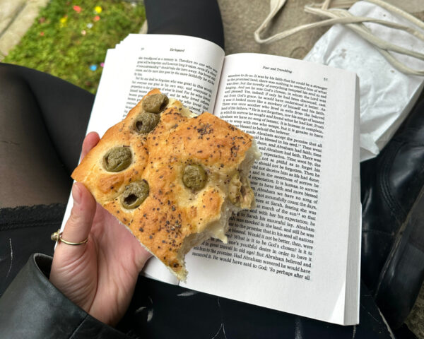Person sitter med en bit focaccia i handen. Focaccian har oliver på sig, och under den ligger en uppslagen bok.