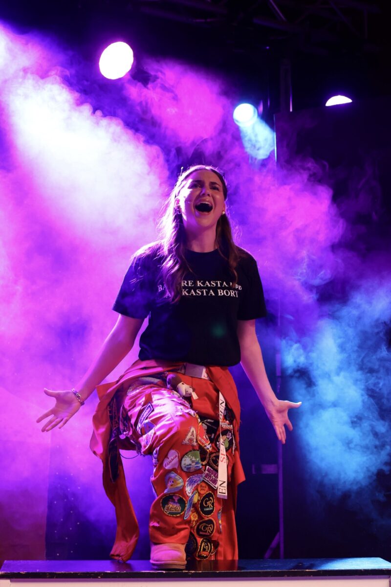 Vilma Ahvenainen klädd i röd halare sjunger på en scen. Scenlamporna bakom henne lyser i lila och blått.