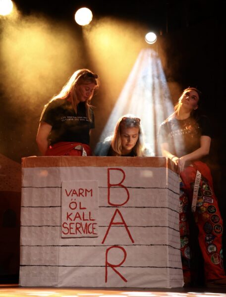 Tre personer klädda i röda halare står vid ett podium på en scen. På podiet står det "BAR" och "varm öl, kall service" med stora, röda bokstäver. ÖLjuset på scenen är gult.