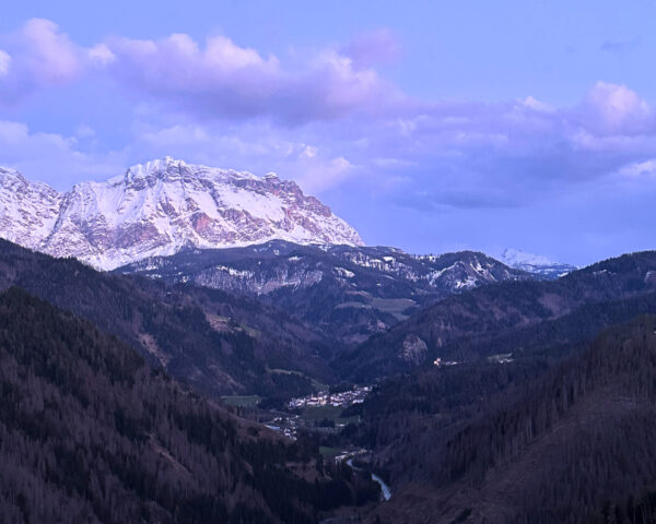 Snötäckta bergstoppar i bakgrunden, i förgrunden en dal med ett litet vattendrag. Molnen är vagt rosa, och hela bilden har ett ljuslila sken.