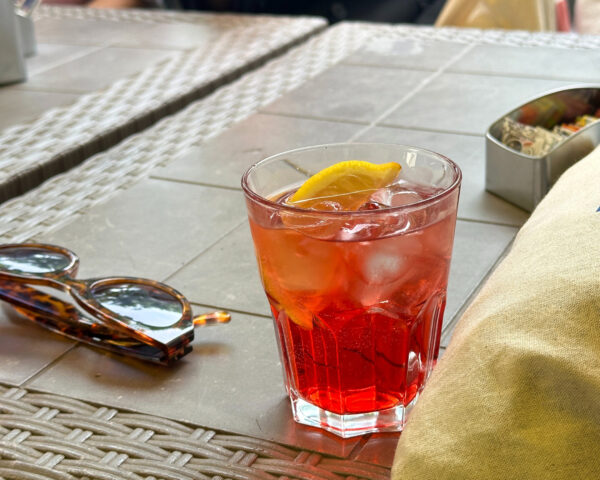 Ett glas står på ett beige bord. I glaset en röd vätska med is och en citronskiva. Bredvid glaset ligger ett par solglasögon och i bakgrunden skymtar ett askfat.