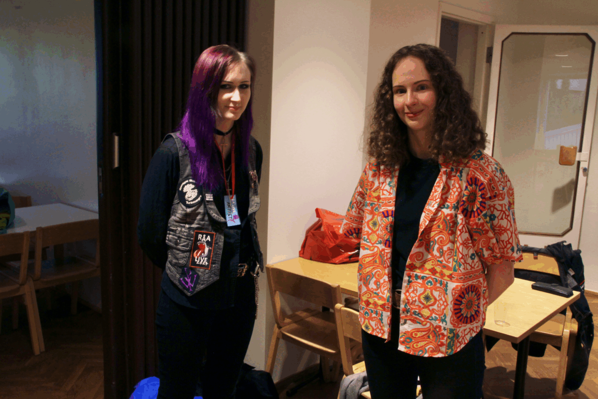 Två personer står och tittar leende in i kameran. Personen till vänster har lila hår, svart ögonsmink och är klädd i svart. Personen till höger har brunt, lockigt hår och en orange-mönstrad skjorta.