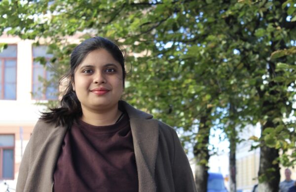 Priya Chakravartty i halvkroppsbild. Hon tittar in i kameran och ler lite. Hon har håret i hästsvans och är klädd i en brun kappa. I bakgrunden syns en gul byggnad, träd och människor som går förbi.