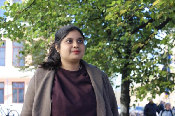 Priya Chakravartty i halvkroppsbild. Hon tittar upp till höger och ler lite. Hon har håret i hästsvans och är klädd i en brun kappa. I bakgrunden syns en gul byggnad, träd och människor som går förbi.