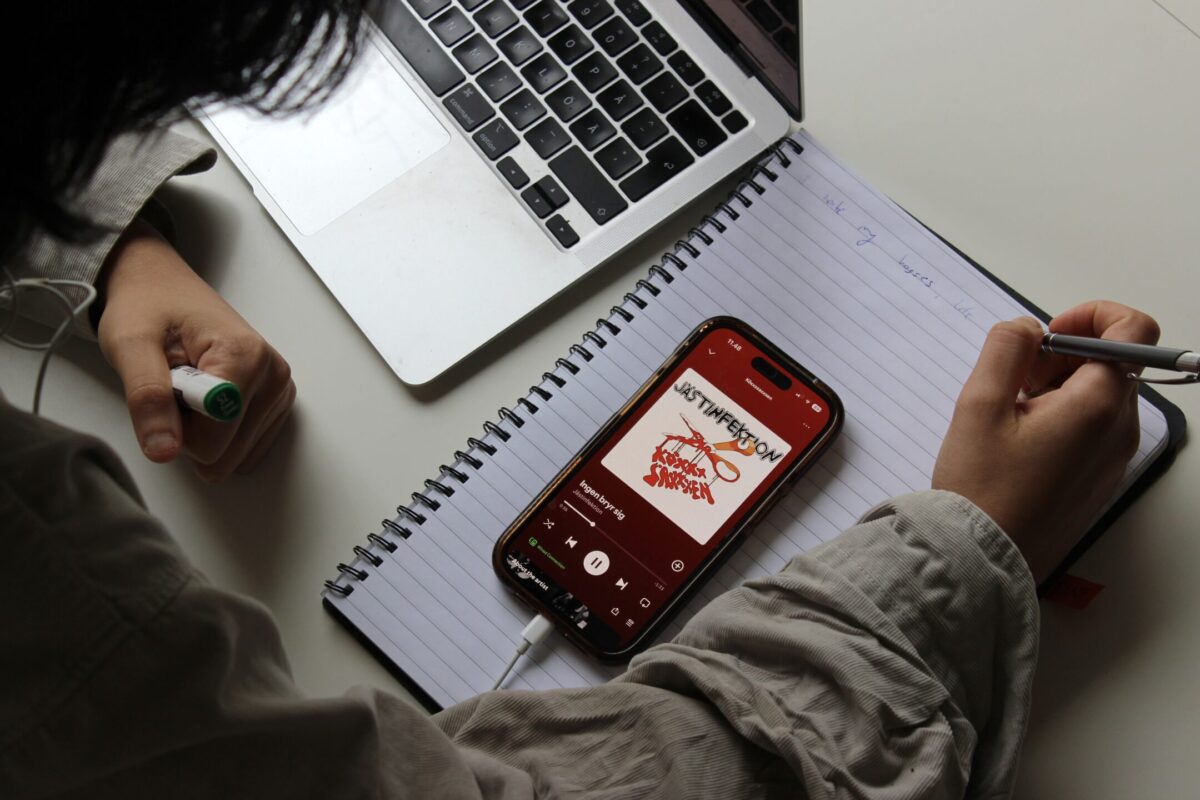 Person skriver i ett häfte. På häftet ligger en mobil som visar att låten "Ingen bryr sig" av Jästinfektion spelar. 