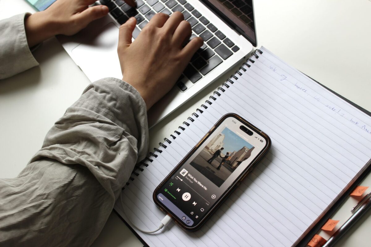 Person skriver på sin laptop. Bredvid laptopen ligger ett häfte. På höftet ligger en telefon, som visar att personen lyssnar på Wish you were here av Pink Floyd.