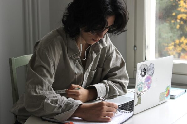 En person sitter vid ett bord. Hen studerar med en öppen laptop och skriver i ett häfte. Hen har trådhörlurar i öronen.