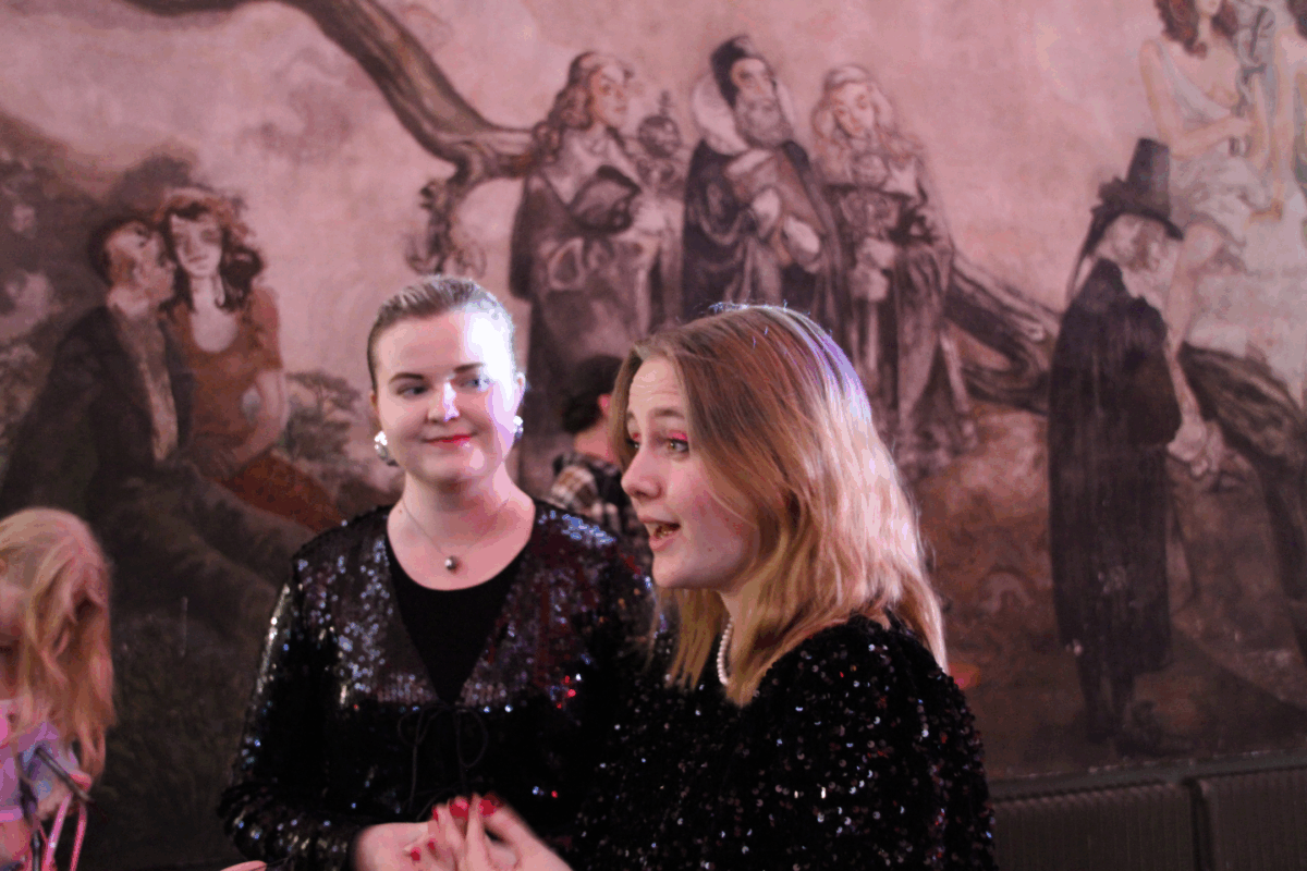 Två personer står framför en vägg med motiv av människor. Personerna framför väggen är klädda i svarta festliga kläder och en av dem har chockrosa ögonskugga.