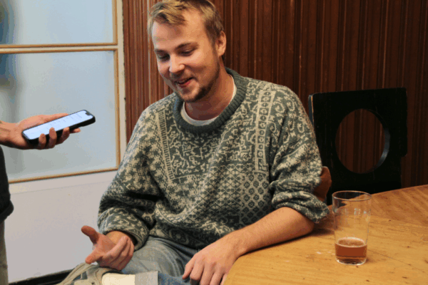 En person iklädd en tröja sitter vid ett bord med ett ölstop som nästan är tomt. Hen ser glad ut och pratar.