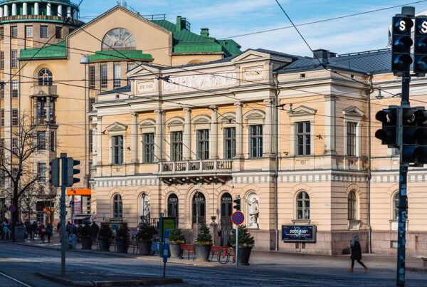 Gult hus med grönt tak i solljuset. I förgrunden flera människor och några trafikljus.