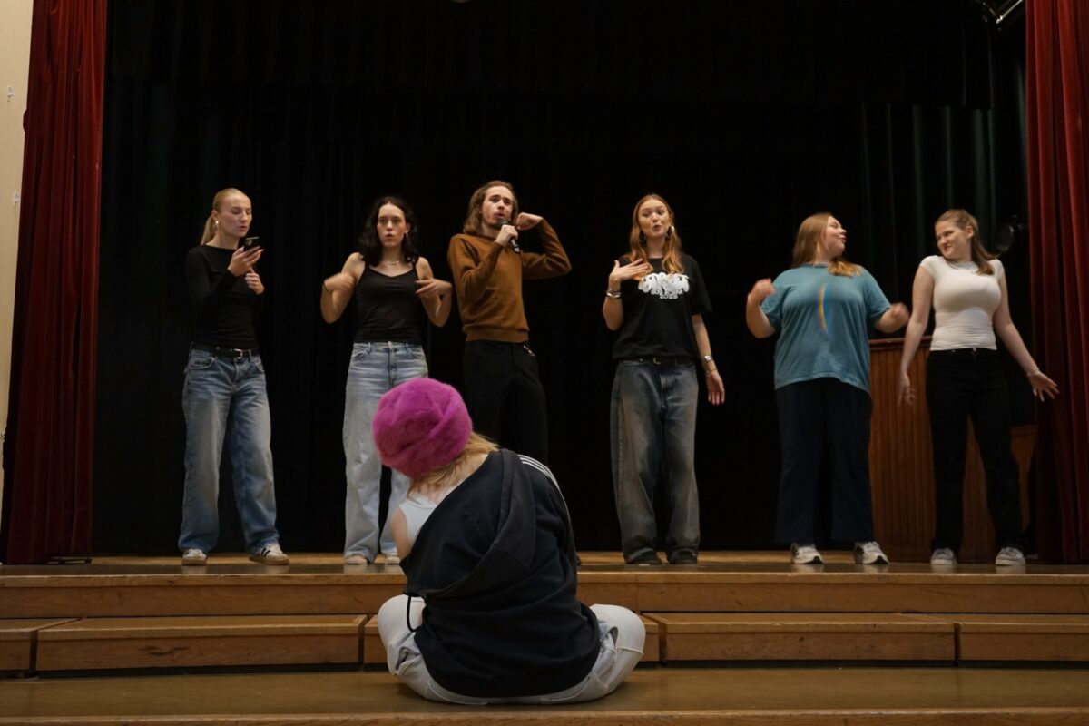 En grupp står på en scen. En person har en mikrofon i handen. Framför dem sitter en annan person med ryggen mot kameran. Hen har på sig en rosa, fluffig hatt och ser ut att gunga från sida till sida i takt med sången.
