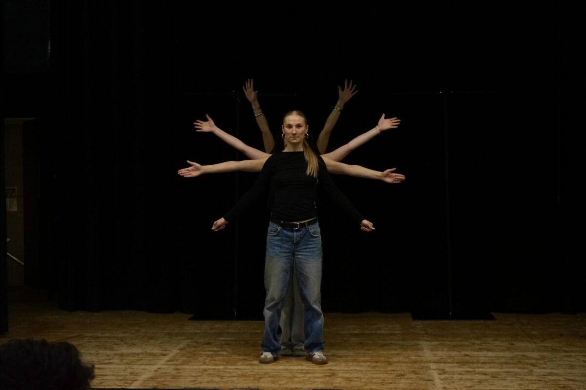 En person står på scen med sina armar nedsträckta. Bakom hen står tre andra personer och sträcker ut sina armar i olika höjd, så att det ser ut som att personen längst fram har en cirkel av armar runt omkring sig.