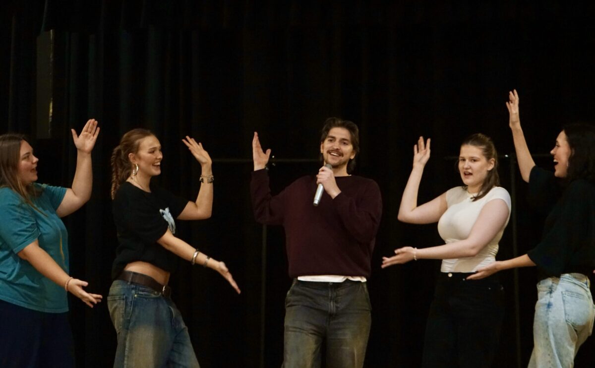 En grupp personer på en scen. Personen i mitten sjunger i en mikrofon och gestikulerar mot sig själv och resten håller ut händerna mot personen i mitten.