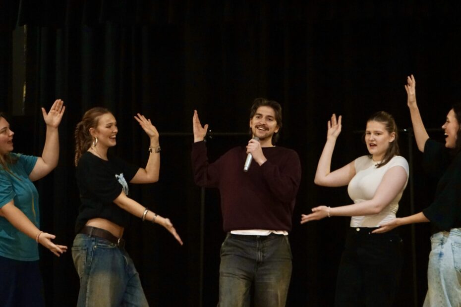 En grupp personer på en scen. Personen i mitten sjunger i en mikrofon och gestikulerar mot sig själv och resten håller ut händerna mot personen i mitten.