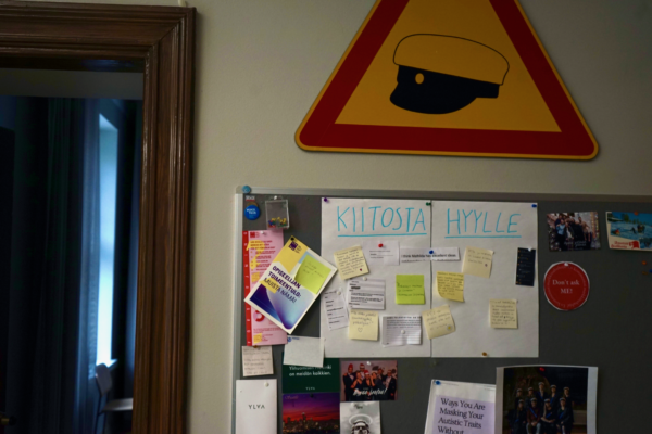 En anslagstavla med flear studentrelaterade annonser och lappar. På en stor lapp i mitten står det "KIITOSTA HYYLLE". Ovanför anslagstavlan hänger en triangelformad trafikskylt. Den är gul med röd kontur och har svarta konturer av en studentmössa i mitten.