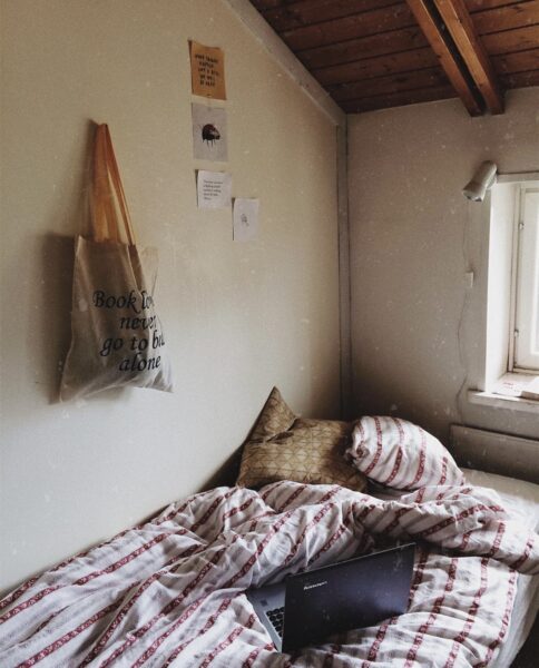 En säng i ett litet rum. På väggen hänger en tygpåse med någonting i. Bredvid tygpåsen en plansch.