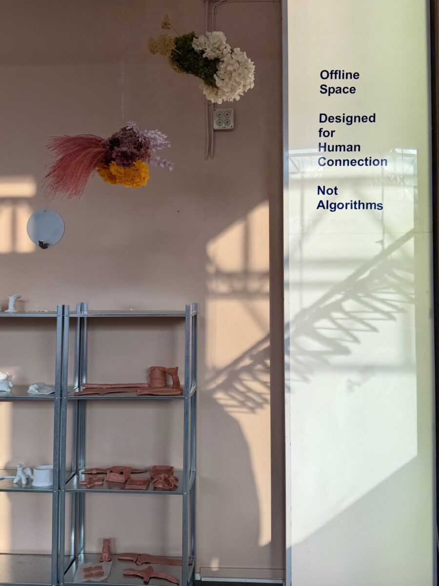 En bild på ett rum. Väggen är gammelrosa och i taket hänger torkade blommor. På e metallhylla längst bak i rummet syns en rad lerskulpturer. I förgrunden syns en skylt med texten "Offline space designed for human connections, not algorithms"
