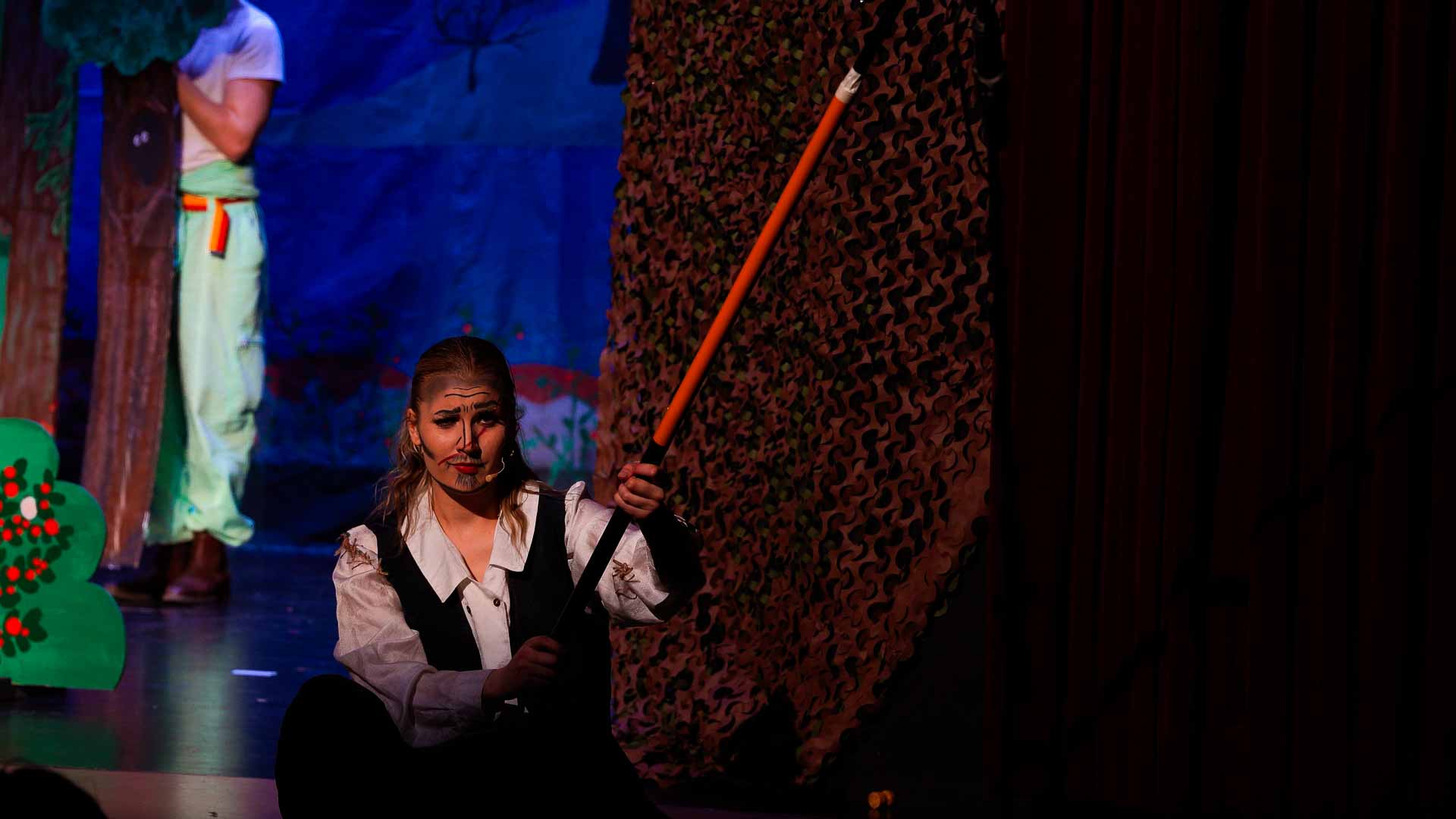 Lemminkäinen står på knä med en käpp riktad mot himlen. Han har ena ögonbrunet höjt och ler smått.