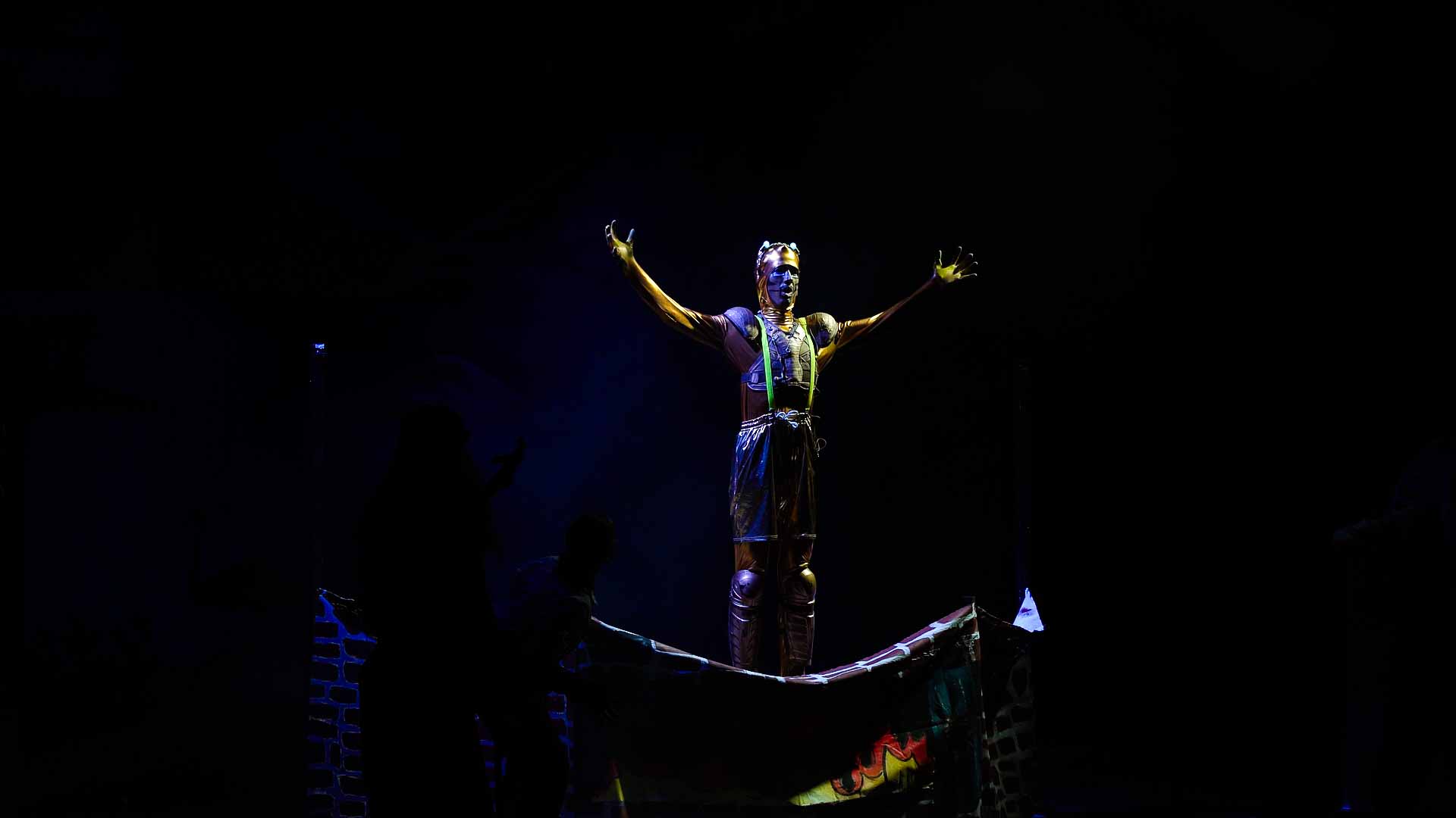 Kvarnen Sampo står på en piedestal i mörkret med armarna utrsträckta.