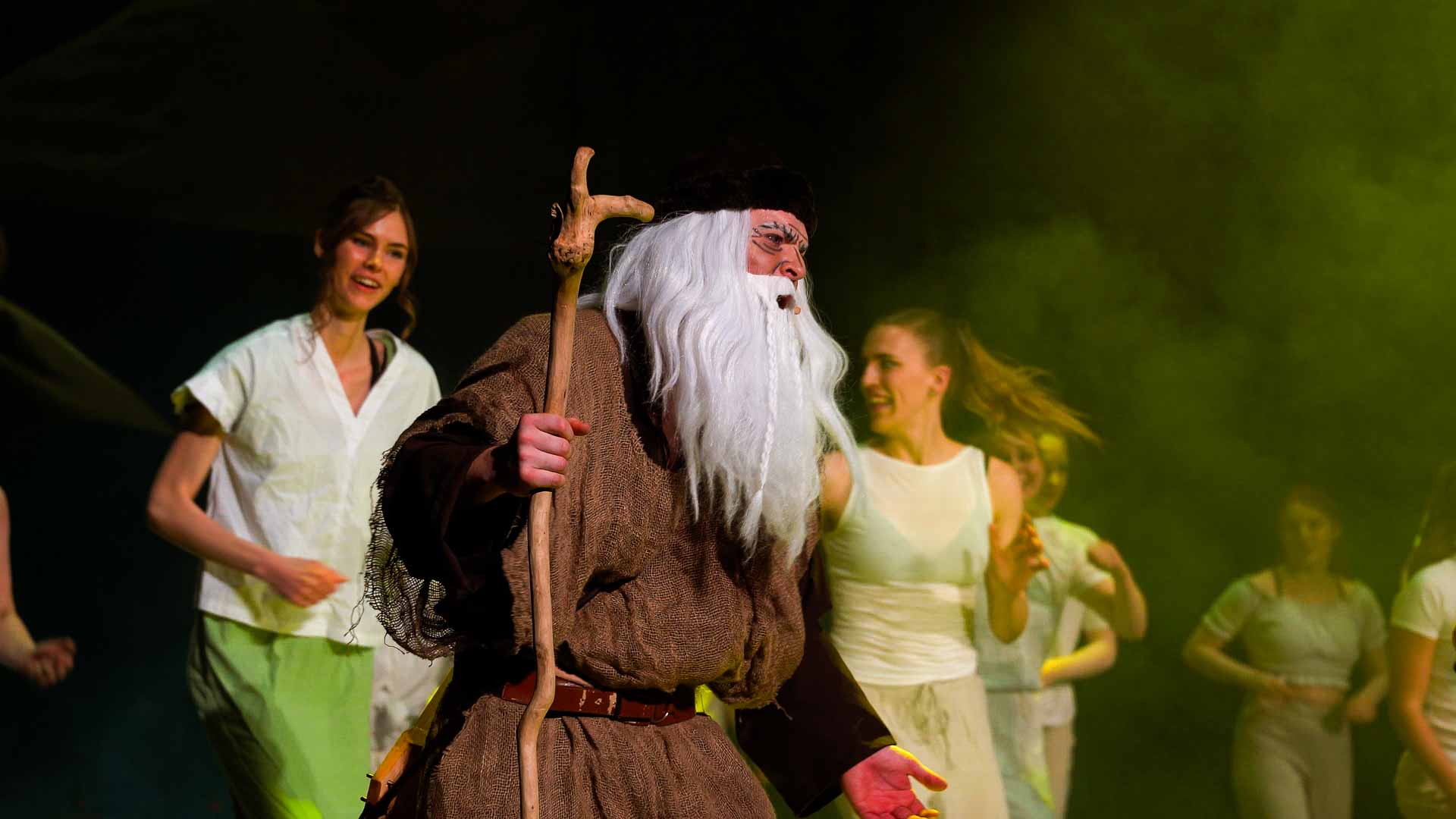 Väinämöinen sjunger. Han håller i en brokig stav, har långt vitt skägg, en brun kläddräkt och en liten svart mössa.