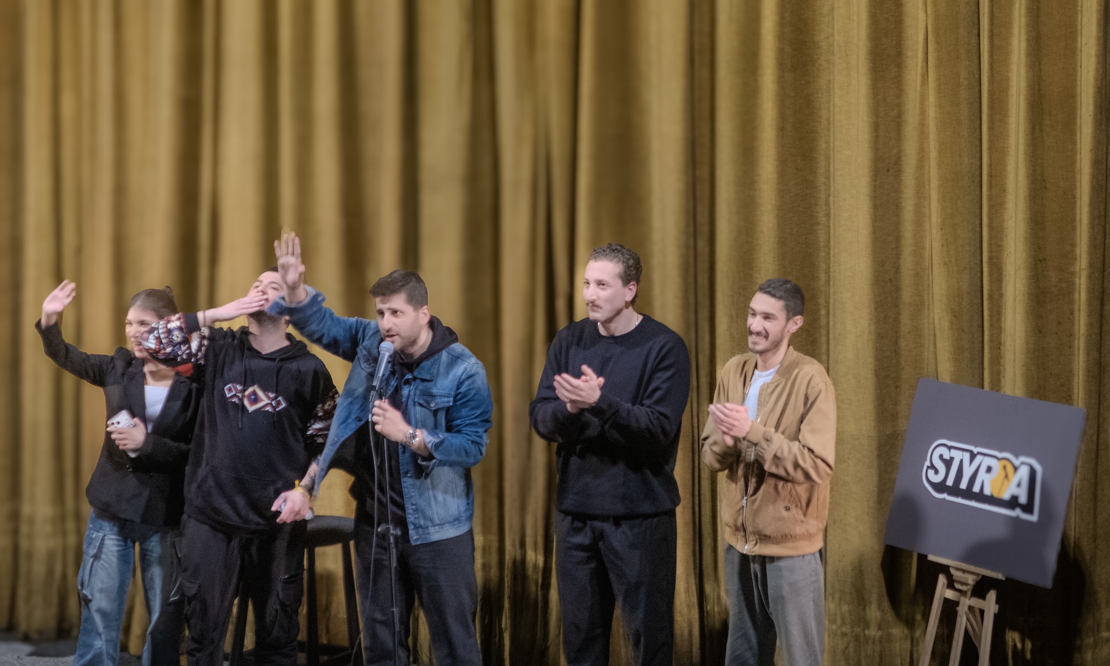 En grupp komiker på en schen. Bredvid dem ett lila plakat med texten "Styria"