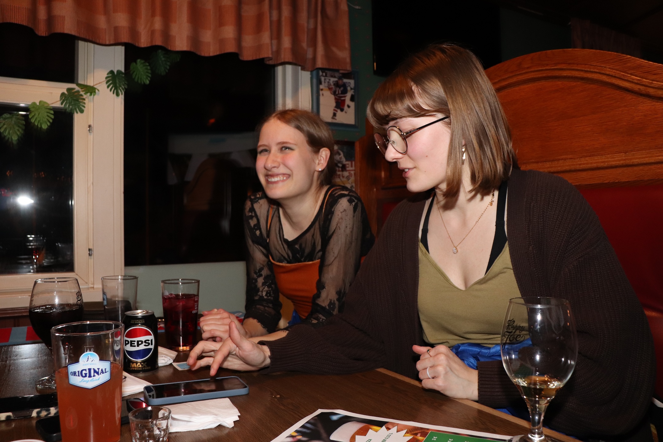 Vanessa Havukoski och Frida Koskinen sitter i ett bås på en pub. Koskinen tittar på sin telefon och Havukoski tittar på något bakom kameran.
